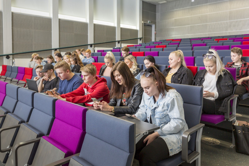Joukko opiskelijoita istuu auditoriossa, osa katsoo kännykkää.