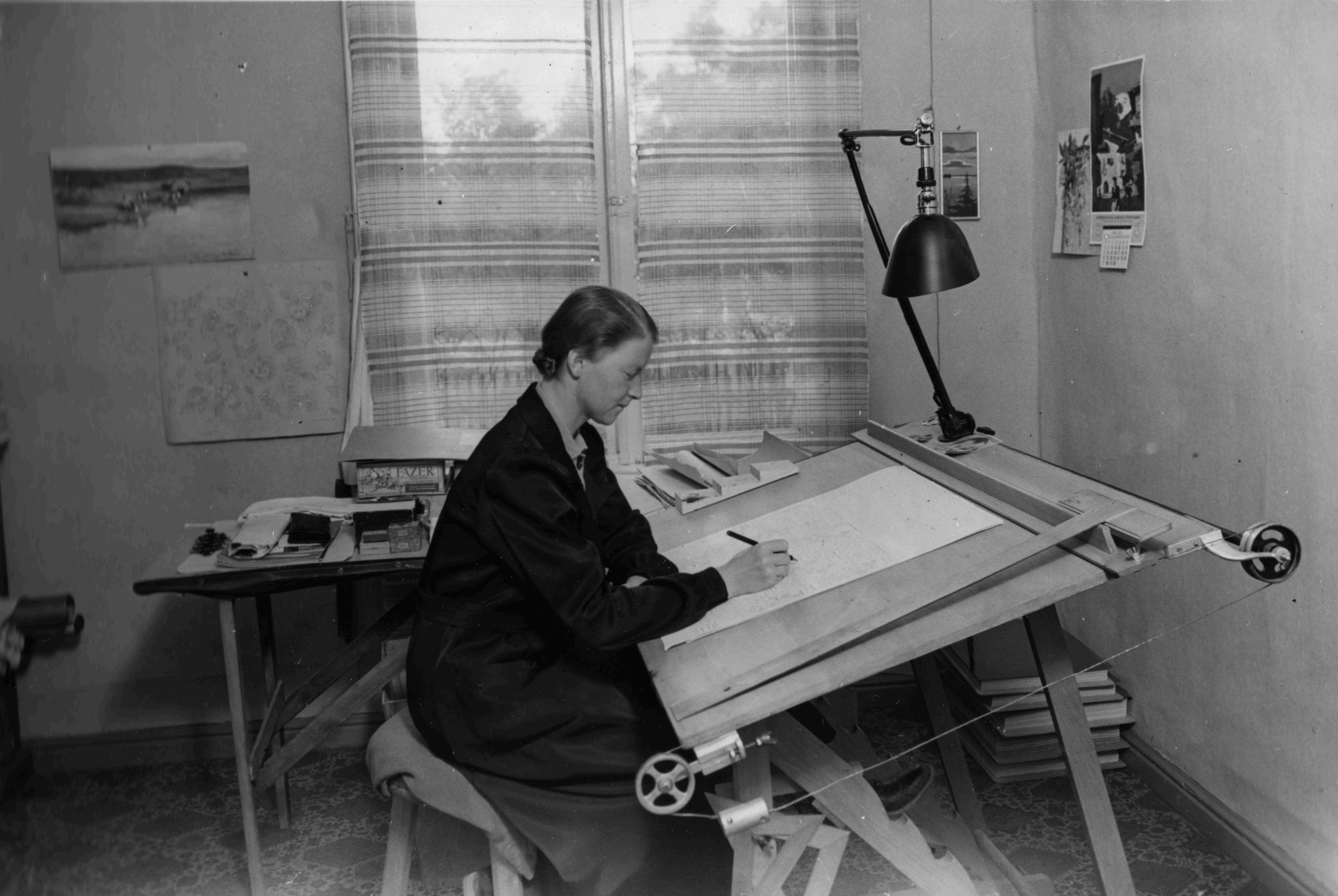 Vanha mustavalkoinen kuva, jossa nainen istuu työpöydän ääressä ja piirtää.