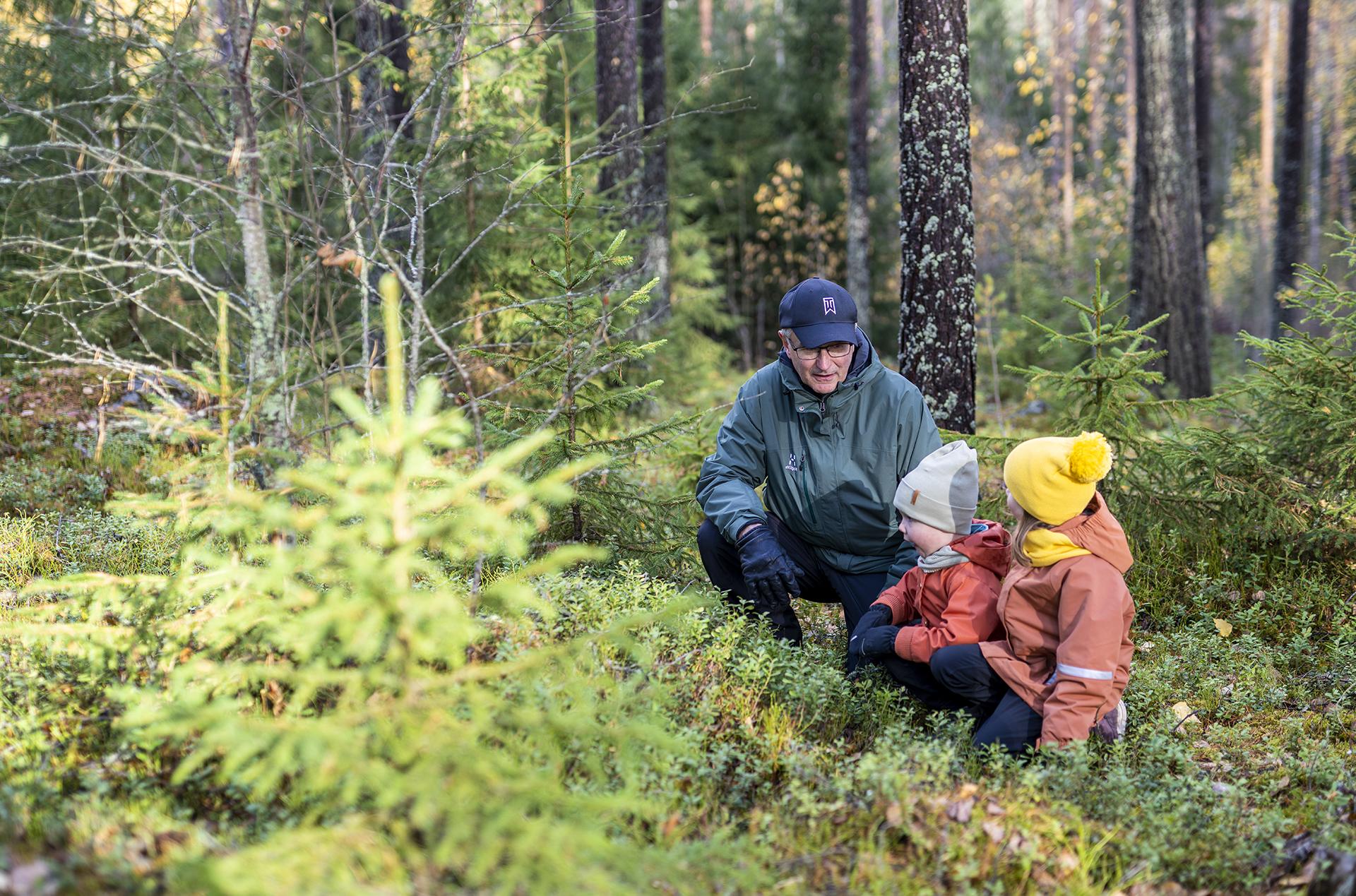 Mies ja kaksi lasta istuvat kyykyssä metsässä ja keskustelevat.